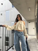 Blusa color crema con escote halter y diseño strapless con hombros descubiertos, mangas largas abullonadas con puños elastizados y corte corto.
