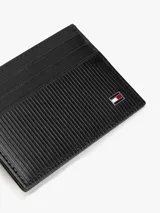 Tarjetero de cuero negro con diseño compacto y plano. Presenta múltiples ranuras para tarjetas y el logo metálico de la bandera de Tommy Hilfiger en la esquina inferior derecha.