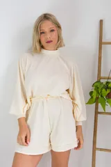 Conjunto de dos piezas en color crema, compuesto por un buzo de cuello alto, mangas largas abullonadas y ajuste en la cintura, junto con un short de tiro alto con terminaciones al corte.