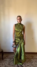 Vestido corto sin mangas con cuello alto, confeccionado en tafeta de color marrón con acabado brillante. Presenta silueta entallada, drapeados frontales y un gran nudo lateral con una larga cola que cae hasta el suelo.