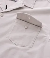 Camisa boxy color crema de algodón texturizado, con cuello inglés, mangas cortas, bolsillos delanteros y lettering bordado.