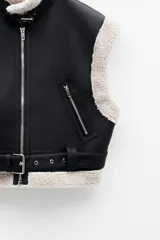 Chaleco biker negro de cuero sintético con forro de borrego beige, cierre frontal metálico, bolsillos laterales con cremallera y cinturón ajustable.