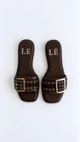 Sandalias planas de dedo cuadrado color marrón, con tira ancha superior decorada con ojalillos metálicos plateados y una gran hebilla dorada.