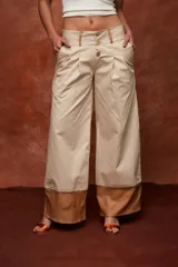 Pantalón ancho de tiro alto color beige, con pliegues frontales y botones visibles en la cintura. El ruedo del pantalón tiene un detalle de terminación en cuero color caramelo.
