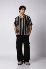 Camisa de manga corta con cuello clásico y cierre frontal de botones. Confeccionada en tejido de punto crochet calado, presenta un patrón de rayas verticales negras sobre fondo crudo.