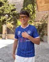 Camiseta de fútbol de manga corta en color azul con diseño estampado abstracto y cuello en V rosado con detalles blancos a juego con los puños. Presenta inscripciones y escudos deportivos en el pecho.