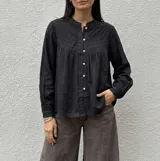 Camisa negra de corte amplio y fluido, con cuello mao, mangas largas y cierre frontal de botones. Presenta un canesú con detalles bordados y calados en la parte superior delantera.