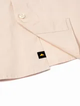 Camisa de manga larga color beige, estilo utilitario, con cuello clásico y cierre frontal de botones. Presenta tres bolsillos de carga en el pecho con solapas y cierre de velcro, además de un detalle de argolla metálica triangular y etiqueta con logo de la marca.