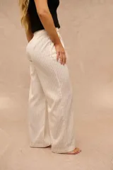 Pantalón de vestir color crudo con rayas diplomáticas negras, de tiro alto, corte sastrero y piernas anchas. Cuenta con bolsillos laterales y cierre frontal con botón carey.