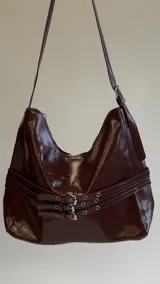 Cartera bandolera color bordó de charol con tiras decorativas cruzadas con hebillas metálicas en el frente y correa regulable.