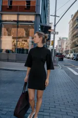 Vestido corto negro de tejido de punto, con cuello redondo alto y mangas anchas a la altura del codo. Presenta un corte entallado en la cintura y cierre con cremallera oculta en la espalda.