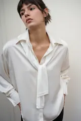 Camisa de manga larga confeccionada en satén de color blanco, con superficie lisa, leve brillo y caída suave. Presenta cuello clásico y cartera frontal.