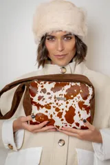 Cartera de mano tipo sobre confeccionada en cuero vacuno con pelo, presentando un diseño estampado con manchas blancas sobre fondo marrón. Posee detalles en gamuza marrón en los laterales y una placa metálica con el logo de la marca en el frente.