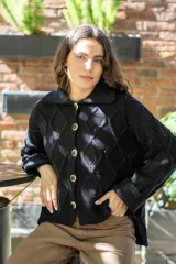 Saco tejido de punto con diseño de ochos y trenzas, color negro. Presenta cuello estilo camisero, cierre frontal con botones tipo joya, mangas holgadas y silueta oversize.