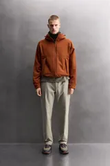Chaqueta de hombre color marrón óxido con textura de pequeños cuadrados en relieve. Presenta cierre frontal de cremallera, capucha ajustable, bolsillos laterales con solapa y puños ajustables. El interior es de forro polar.