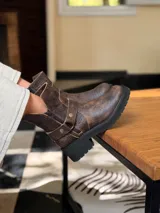 Bota corta de cuero vacuno en color chocolate con efecto desgastado. Presenta una suela track de goma negra, detalle de pulsera desmontable con tachas metálicas y cierre lateral.