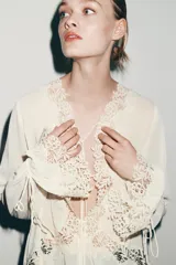 Blusa de escote en pico con manga larga terminada en volantes. Presenta aplicaciones de encaje a tono, cierre frontal con lazadas y bajo asimétrico.