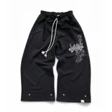 Pantalón de jogging negro de corte baggy, confeccionado en algodón. Presenta una estampa plastisol con bordado decorativo en la pierna izquierda, cintura elástica con cordón largo ajustable y botones en el ruedo para regular la botamanga.