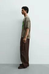 Camiseta de corte relajado con cuello redondo y manga corta. Presenta un efecto de lavado desgastado en tonos marrones y un estampado frontal de una cabeza de alienígena en color verde vibrante.