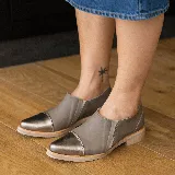 Zapato tipo mocasín de cuero vacuno en tono gris topo, con puntera reforzada en cuero metalizado color bronce. Presenta un diseño de corte lateral y suela plana con pequeña elevación en el talón.