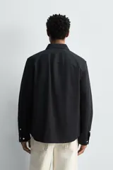Sobrecamisa de corte relajado confeccionada en tejido ligero de algodón color negro. Presenta cuello de solapa, manga larga con puños abotonados y dos bolsillos de plastrón con solapa en el pecho. Cuenta con cierre frontal de botones a contraste y aberturas laterales en el bajo.