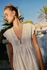 Vestido largo color crudo, sin mangas, con escote en V pronunciado, canesú superior de tejido de punto con botones frontales y lazo para atar en el cuello, y falda fluida con detalle de bordado en el bajo.