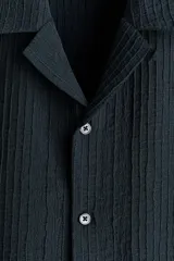 Camisa de manga corta color negro con textura acanalada, cuello tipo resort, cierre frontal con botones y dos bolsillos aplicados al frente.