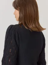 Buzo de lana color beige con mangas abullonadas de tejido calado con diseño de rombos. Presenta un cuello redondo y un detalle de flor negra de tela con cintas colgantes en el escote.