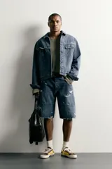 Bermuda baggy fit confeccionada en denim de algodón con lavado azul y efecto rotos en toda la prenda. Presenta diseño de cinco bolsillos, detalles de costuras en las perneras, bajo acabado irregular y cierre frontal con cremallera y botón.