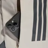 Mochila Adidas Primelift de color beige con detalles en gris oscuro, incluyendo correas de compresión laterales y refuerzo en la base. Presenta el logo de la marca y las icónicas tres tiras estampadas en la parte frontal.