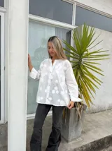Camisa blanca de corte holgado con cuello mao y cierre frontal de botones. Presenta un diseño bordado con motivos florales en toda la prenda y mangas largas con puños amplios.