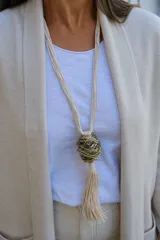 Collar largo con cordones de algodón color crema, que presenta un dije central esférico formado por hilos metálicos entrelazados en tonos dorados y plateados, finalizando con una borla de flecos de algodón.