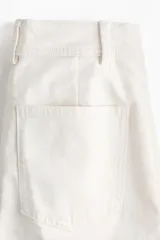 Pantalón de sarga color blanco de corte acampanado y tiro alto, con bolsillos delanteros de parche con solapa y botones oscuros, y pliegues marcados en el centro de las piernas.