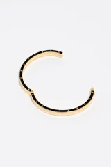 Pulsera rígida tipo brazalete de color dorado con un detalle circular de esmalte negro que presenta el logo de la marca. Cuenta con un cierre de bisagra para mayor seguridad.