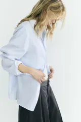 Camisa oversize de algodón con estampado de rayas finas, cuello solapa, escote en pico y manga larga con puños abotonados. Presenta un diseño con pliegues laterales y cierre frontal con botones.