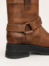 Bota de punta cuadrada realizada en cuero encerado color marrón, con hebillas en la caña, herrajes de níquel y fondo de suela negro.