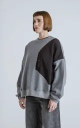Buzo de cuello redondo con diseño oversize y corte crop. Confeccionado en frisa de algodón color gris melange, presenta un recorte asimétrico en color negro en microfibra con un bolsillo frontal funcional con cierre a presión.