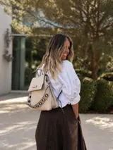Mochila tipo cartera de cuero sintético color beige, con tapa frontal, compartimentos internos y detalles de ojales metálicos en las correas.