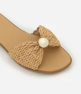 Sandalia plana de color beige con tira ancha confeccionada en rafia trenzada con diseño de nudo. Presenta un detalle decorativo de esfera perlada en el centro, punta redondeada y suela fina de 1 cm.