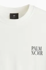 Remera de algodón blanca de corte holgado, con cuello redondo y estampa pequeña con el texto "PALM NOIR" en el pecho.