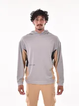 Sudadera con capucha (canguro) color gris claro, con bloques de color en negro y beige en los laterales y mangas. Presenta un pequeño texto estampado en el frente.