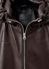 Chaqueta de cuero de napa marrón oscuro, de diseño oversize, con capucha ajustable, cierre frontal de cremallera, bolsillos laterales y bajo ajustable con cordón.