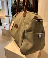 Bolso amplio de gamuza azul con manijas largas de cuero marrón. Cuenta con bolsillos laterales, uno con clip, cierre posterior y un bolsillo interno amplio. Presenta detalle de bandera y logo bordado en los laterales.