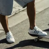 Championes adidas modelo Puig Indoor, de diseño bajo en color blanco con las icónicas tres tiras en azul claro y detalles en gamuza gris en el talón y la zona de los cordones. Presenta el nombre del modelo en dorado en el lateral.