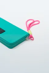 Billetera rectangular con cierre, color celeste pálido, con parche frontal negro que lleva las letras "CNJ" en relieve. Incluye un llavero de muñequera con cordón trenzado en rosa fuerte y verde lima.