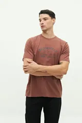 Remera marrón de manga corta con cuello redondo y estampado frontal con el texto "BROOKSFIELD The Emporium of Postmodern Activity No 1001 TORINO | 1971".