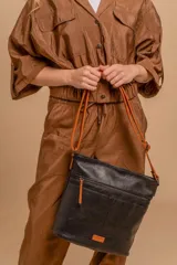 Bandolera mediana de cuero sintético negro con detalles en color naranja. Presenta un bolsillo frontal con cierre, correa regulable y logo de la marca en una etiqueta naranja en la parte inferior.