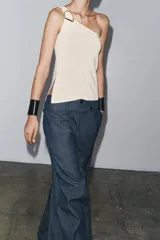 Top blanco de punto con escote asimétrico y aplique metálico dorado en el hombro.