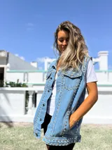 Chaleco de jean azul claro con diseño sin mangas, cuello clásico y cierre frontal con botones metálicos. Presenta una profusión de tachas plateadas distribuidas a lo largo de las costuras frontales y los bolsillos del pecho.