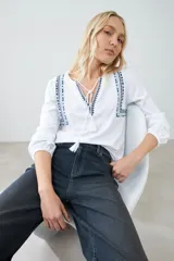 Remera blanca de manga larga con detalles bordados en tonos azules y verdes en el pecho y hombros. Presenta un escote en V con cordones ajustables terminados en borlas y puños elásticos.